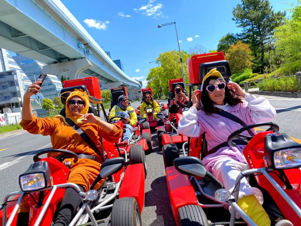 Group of people in colorful animal onesies riding red go-karts on a sunny city street, taking a selfie together under a blue sky.