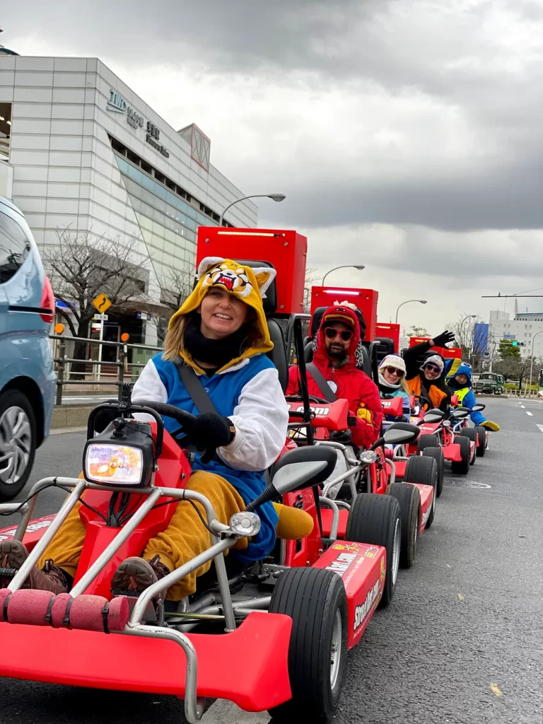 IMG_2602 - StreetKart People riding red pedal go-karts in a parade down a city street, front rider wearing a tiger-hooded onesie and gloves, smiling at the camera under a cloudy sky