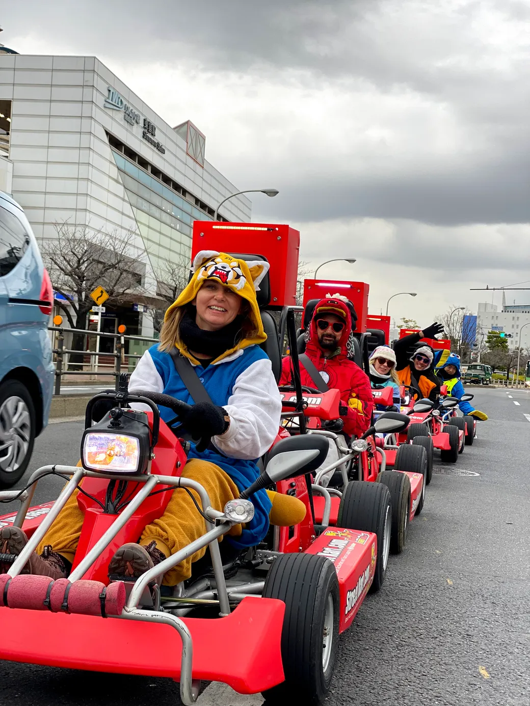 People riding red pedal go-karts in a parade down a city street, front rider wearing a tiger-hooded onesie and gloves, smiling at the camera under a cloudy sky