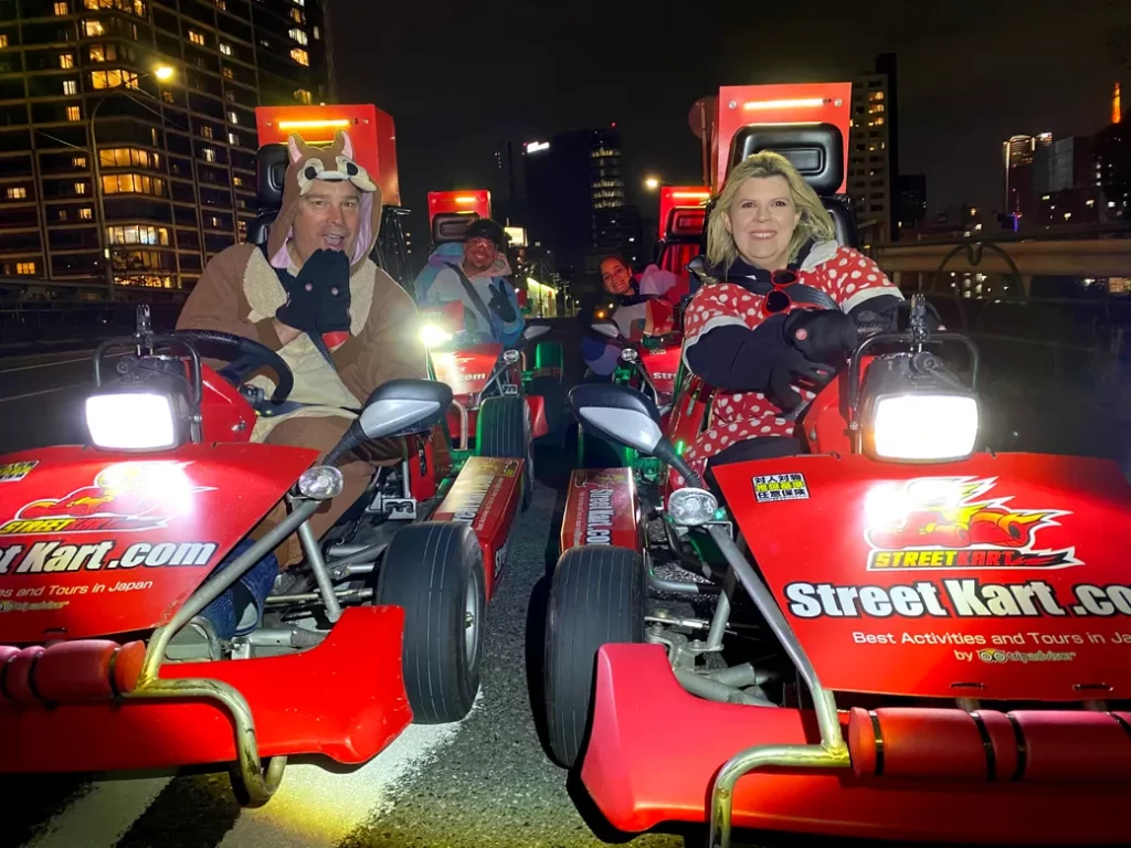 Group of people driving bright red go-karts at night on a city street, wearing costumes and smiling at the camera.
